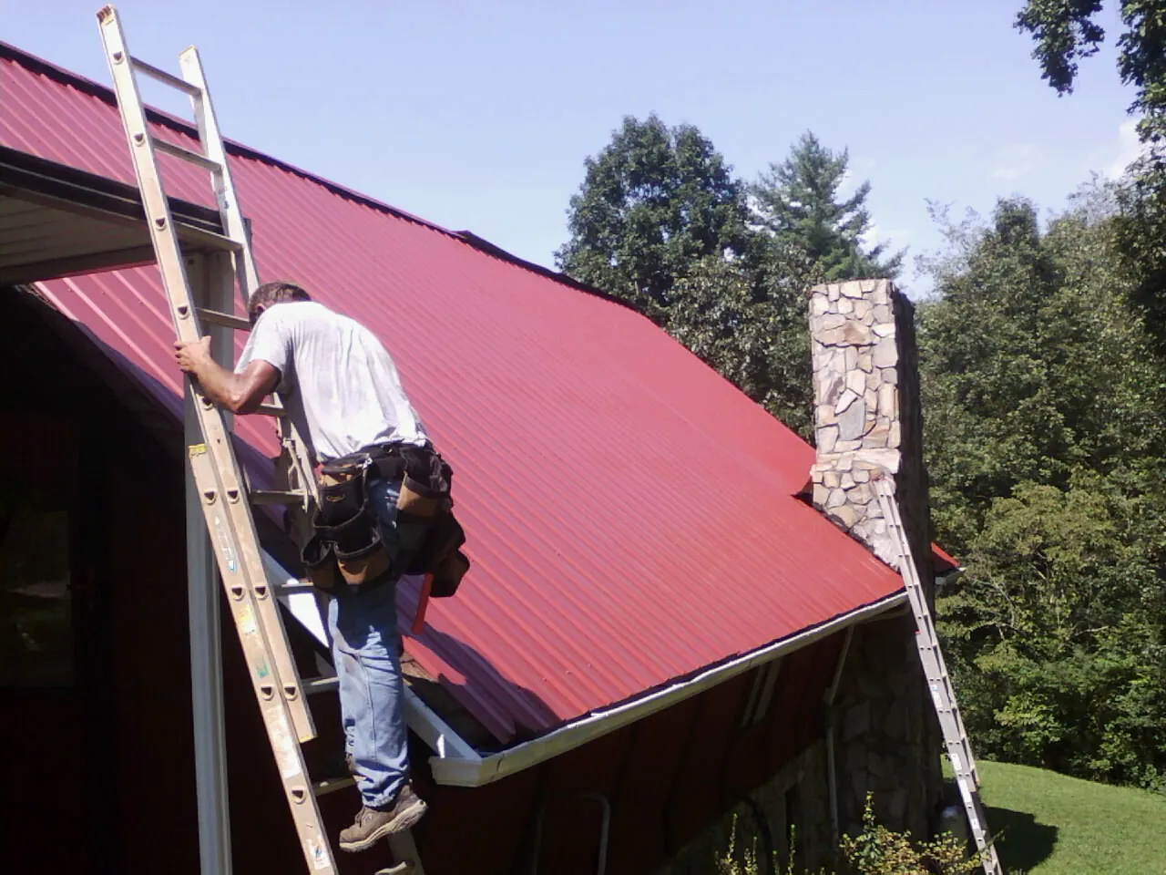 Roofer on a ladder beside a red metal roof and stone chimney — Soffit & Fascia Repair by Roofing Brothers in Tennessee